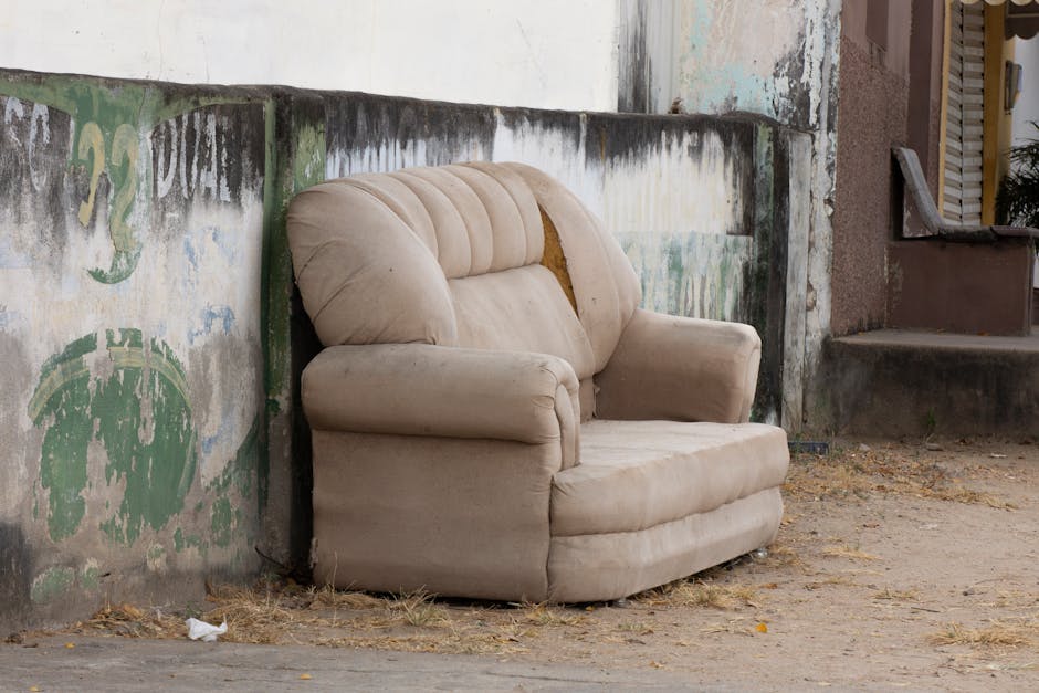 A beige upholstered armchair with a torn and worn fabric exposing yellowish foam padding is positioned on a dirt ground outdoors, next to a weathered concrete wall with peeling paint and faded green and blue graffiti. The armchair appears to be part of a house removal or disposal process, with no cushions or packaging materials visible. It is located adjacent to a doorway with a step leading into a building, and the background includes a staircase and a small section of a window or shutter. The lighting is natural daylight, and the setting suggests an outdoor area where furniture is being prepared for removal or transport, supported by the presence of a company like Man with Van Silvertown, known for house removals and rubbish clearance services.