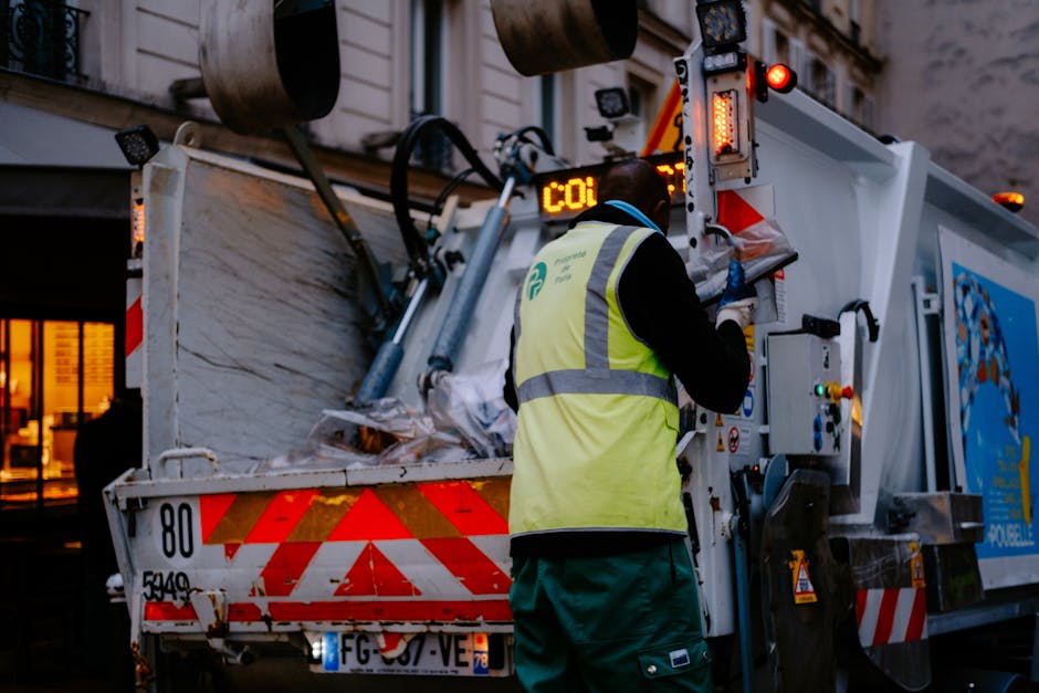 A worker wearing a yellow high-visibility vest and green trousers is operating a waste collection vehicle on a street at dusk, loading and managing large bulky waste items such as furniture and household appliances. The truck's open rear compartment contains a mix of cardboard boxes, wooden furniture pieces, and plastic-wrapped items, with some materials stacked and secured inside. The worker is focused on the control panel, possibly by pressing buttons or adjusting settings related to waste disposal or collection. The vehicle is positioned near a pavement, with a nearby building visible in the background having lit windows, indicating an urban environment. The scene depicts a professional waste removal process, which may be related to house clearance or bulky item disposal services provided by Man with Van Silvertown as part of their house removals and transport offerings that support efficient relocation logistics.