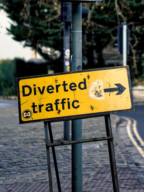 A yellow rectangular road sign with black text reading 'Diverted traffic' and an arrow pointing to the right, mounted on a metal stand positioned on a cobbled pavement roadside. The sign shows some dirt and wear, indicating it has been exposed to the elements. Behind the sign, there is a grey metal pole that supports the sign, and further in the background, there are blurred trees, a road, and a curb with a double yellow line. This scene is related to road closure signage, which may be relevant to house relocation projects involving moving logistics and road arrangements, as provided by Man with Van Silvertown.