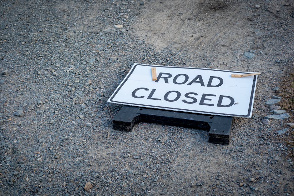 A rectangular portable road closed sign placed on the gravel surface outside, with the words 'ROAD CLOSED' printed in bold black letters on a white background. The sign is supported by a black plastic frame and secured with two wooden clothespins at the top corners. The surrounding ground consists of uneven grey and brown gravel, indicating an outdoor setting likely near a residential or commercial property. This scene depicts preparations for a home relocation or furniture transport, with the sign indicating restricted access to facilitate moving logistics. The sign’s placement suggests it is temporarily used to inform pedestrians and vehicles about road closures, possibly related to a house removal process managed by Man with Van Silvertown, underscoring the importance of planning in moving services involving the loading or unloading of furniture and packing materials.