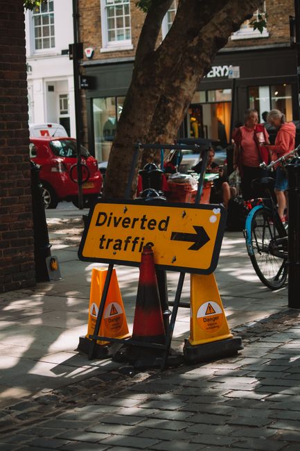 A yellow rectangular road sign with black text reading 'Diverted traffic' and an arrow pointing to the right, mounted on a metal stand positioned on a cobbled pavement roadside. The sign shows some dirt and wear, indicating it has been exposed to the elements. Behind the sign, there is a grey metal pole that supports the sign, and further in the background, there are blurred trees, a road, and a curb with a double yellow line. This scene is related to road closure signage, which may be relevant to house relocation projects involving moving logistics and road arrangements, as provided by Man with Van Silvertown.