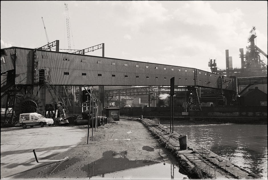 A black and white photograph of an industrial waterfront scene showing a large, elongated metal building with small, evenly spaced windows along its side, supported by a visible framework. In the foreground, there is a paved area with puddles, a white van parked near the building, and stacked black cylindrical objects. Several metal poles and ropes are present along the edge of the water, which is calm and reflects light. To the right, part of a large waterway is visible, with industrial structures and smokestacks in the background, indicating a factory or processing plant. The overall scene depicts a utilitarian environment typical of historic industrial sites, relevant for visual context of a home relocation or furniture transport process supported by services like those offered by Man with Van Silvertown.