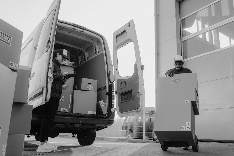 Two removal workers are engaged in a home relocation process during daylight hours, loading cardboard boxes into a parked white van with its side door open. The woman, wearing a cap and dark clothing, is inside the van and appears to be scanning or organizing the boxes, which are securely packed and labeled, while standing on a paved surface. Outside the van, a man wearing a hard hat and dark clothing is pushing a large wheeled container, likely for transporting additional packing materials or smaller items. The background includes a building with large windows and a fence, indicating an industrial or commercial area. The lighting is natural, illuminating the scene clearly, with visible equipment such as blankets or straps that may be used to secure furniture during transport. This scene highlights typical aspects of furniture transport and packing processes involved in house removals, as handled by companies like Man with Van Silvertown.