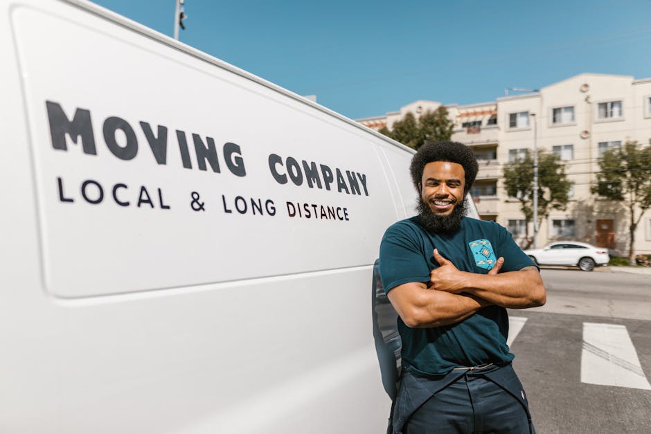A smiling man with an Afro hairstyle and a full beard leans casually against the side of a white moving van on a residential street. He wears a navy blue T-shirt with a small logo on the chest and dark trousers, with arms crossed as he poses for the camera. The van displays bold black text that reads 'MOVING COMPANY' with smaller text underneath stating 'LOCAL & LONG DISTANCE.' In the background, there are modern white apartment buildings with multiple windows, balconies, and some trees indicating an urban environment. The street has parked cars and a pedestrian crossing, with clear daylight and a blue sky overhead. The scene appears to be part of a home relocation or furniture transport process, with the man representing a representative from Man with Van Silvertown during a local or long-distance moving service, aligned with the context of house removals and packing and moving logistics.