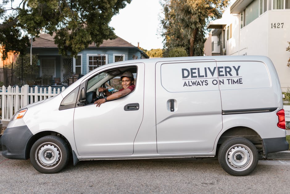 A silver delivery van parked on a residential street during daytime, with a smiling male driver sitting in the driver's seat, leaning out of the window. The van displays the words 'DELIVERY ALWAYS ON TIME' in bold black letters on the side panel. Inside the van, visible packing materials include cardboard boxes and plastic wrapping, suggesting a home relocation or moving process. The background features a white picket fence, a light blue house with multiple windows, and a tree with green foliage, indicating a suburban environment. The setting is well-lit with natural sunlight, and the van appears ready for furniture transport or packing and moving services associated with house removals, managed by Man with Van Silvertown.