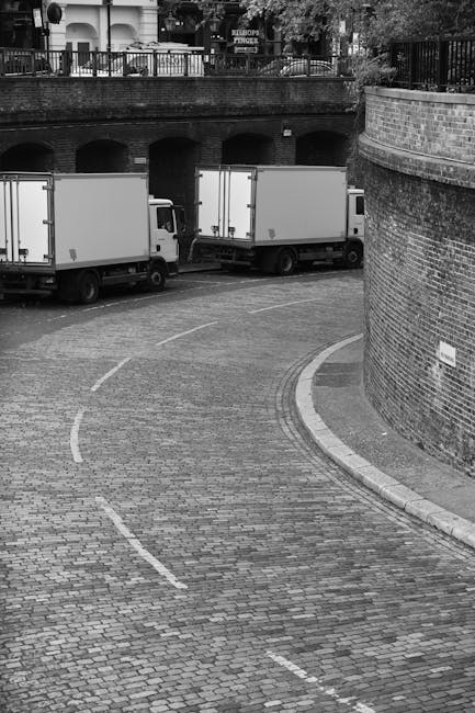 A narrow cobbled street with a gentle curve, showing two white box trucks parked parallel to a brick wall on the right side, which partially encloses an underground parking or loading area. The trucks are positioned close to the curb, with the rear of one vehicle visible, including closed cargo doors. In the background, there is a raised area with a metal railing and a row of trees, suggesting an outdoor residential or commercial setting. The sidewalk runs along the brick wall, and the pavement appears clean and well-maintained. The scene is in black and white, emphasizing the textured cobblestone surface and the structural elements of the loading zone. This setup illustrates the typical environment for home relocation or furniture transport, where efficient parking and loading are essential for moving services such as those offered by Man with Van Silvertown.