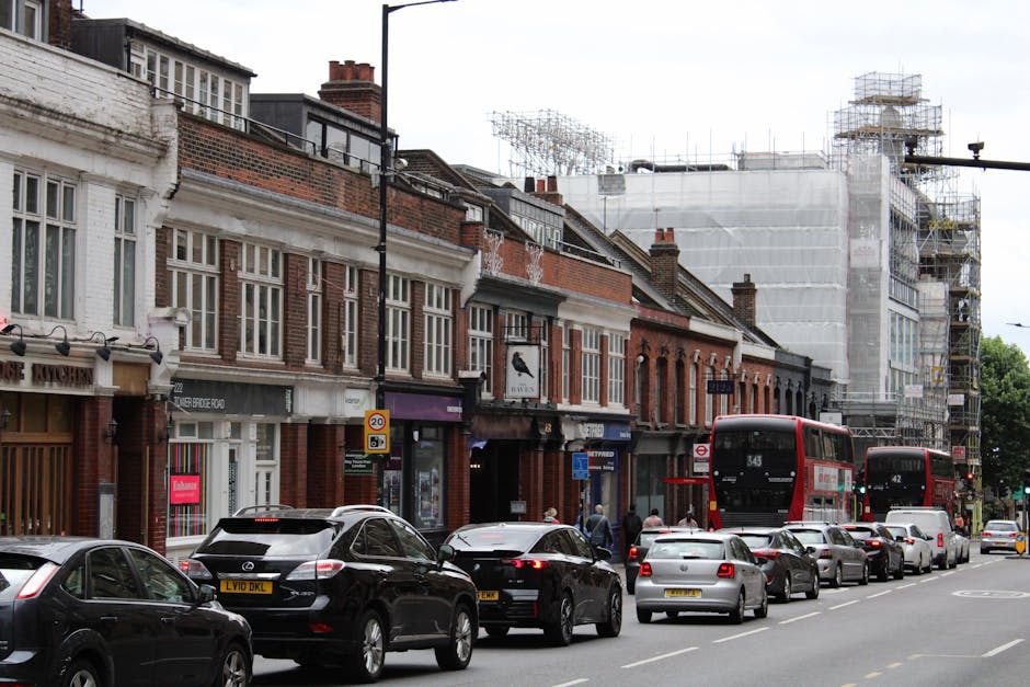 The image depicts a busy urban street scene on North Woolwich Road during daytime, featuring a line of parked cars along the pavement, including sedans and SUVs, facing away from the camera. Two red double-decker buses are seen moving along the street, with the closest bus displaying the route number 343. In the background, there are several brick terraced houses with white-framed sash windows, some with shopfronts on the ground level, and one with a sign for a hairdresser. To the right, a building under construction is covered in scaffolding and white protective sheeting, indicating ongoing building work. The scene includes pedestrians walking along the sidewalk, some near a small group waiting at a bus stop. Man with Van Silvertown's moving van, a white vehicle with branding, is visible parked on the street or nearby, supporting local home relocation and furniture transport services, as part of the home packing and loading process typical of house removals in the area. The overall environment is well-lit, with overcast skies and urban activity centered around traffic management and moving logistics.