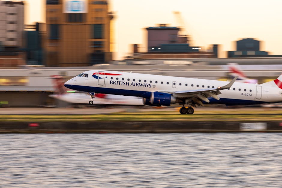 A British Airways commercial airplane with a white fuselage, blue tail fin, and red accents is in mid-air during the takeoff or landing process at an airport, close to the ground near a body of water. The aircraft's landing gear is extended, and the background features blurred images of airport buildings and other planes, indicating motion and speed. The scene is captured during daylight hours with soft natural lighting. This image illustrates the logistics involved in home relocation and the importance of efficient transportation for moving and delivery services, similar to those offered by Man with Van Silvertown in the context of house removals and furniture transport from the Royal Albert Dock to London City Airport.