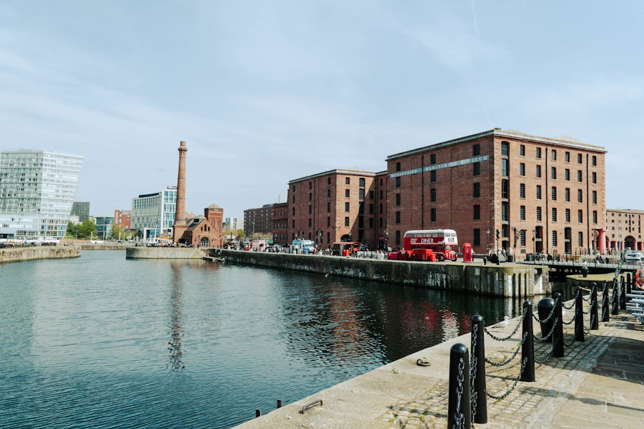 A cityscape view of the Royal Albert Dock area with a calm waterway in the foreground, lined with a stone walkway featuring black bollards connected by chains. Several large, red brick warehouse-style buildings are situated along the dock, with some displaying windows and service doors. A vintage red double-decker bus is parked near the buildings, and some cars are visible along the street, indicating active loading or moving activity. A tall industrial chimney extends upward behind the buildings, contrasting with modern office structures visible further in the background. The scene is illuminated by daylight under a partly cloudy sky, capturing the environment where house removals or furniture transport might take place, as part of a home relocation or moving service coordinated by Man with Van Silvertown.