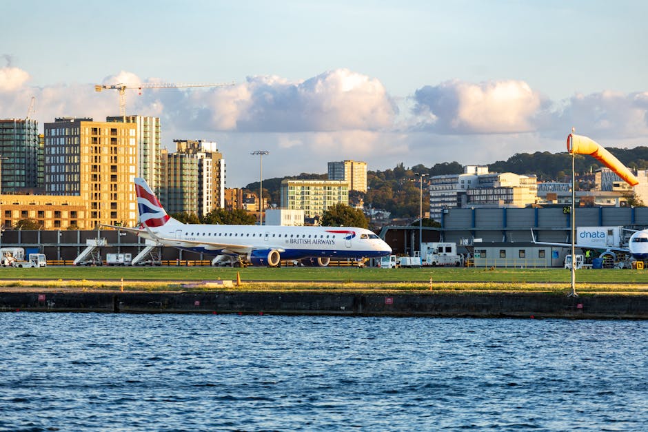 A British Airways commercial airplane with a white fuselage, blue tail fin, and red accents is in mid-air during the takeoff or landing process at an airport, close to the ground near a body of water. The aircraft's landing gear is extended, and the background features blurred images of airport buildings and other planes, indicating motion and speed. The scene is captured during daylight hours with soft natural lighting. This image illustrates the logistics involved in home relocation and the importance of efficient transportation for moving and delivery services, similar to those offered by Man with Van Silvertown in the context of house removals and furniture transport from the Royal Albert Dock to London City Airport.
