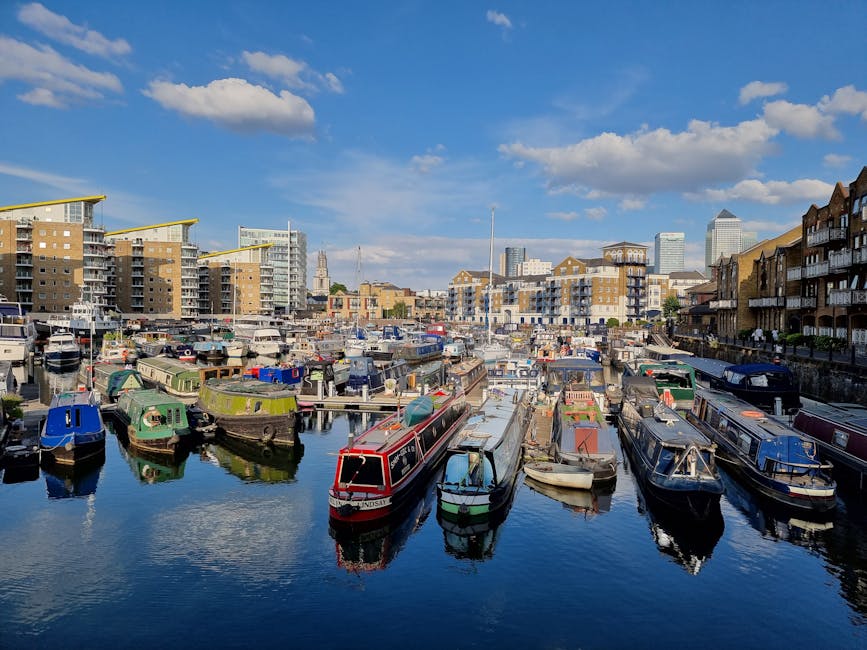 A marina with multiple moored narrowboats and small sailing vessels, anchored along the edge of calm water under a partly cloudy sky. Behind the boats, there are modern residential buildings with balconies and rooftop extensions, alongside taller office or apartment blocks in the distance. The scene depicts the waterfront area in Silvertown, with a clear view of the urban skyline and surrounding structures. The boats include various colors such as green, blue, and red, and are secured with ropes. The water reflects the boats and buildings, creating a mirror-like effect. Nearby, a paved walkway and small dock area are visible, suggesting a location used for boat access and home relocation activities. This setting provides a typical scene for shipping, loading, or preparing for home removals or furniture transport through the area, supported by ongoing moving services like those offered by Man with Van Silvertown.
