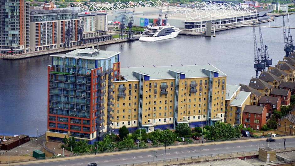 An aerial view of a multi-storey residential building with yellow and blue exterior walls, situated alongside a river in Silvertown, London. In the foreground, a road runs parallel to the building, with parked cars and greenery along the pavement. Behind the building, there is a waterfront area with a white yacht docked near a modern sports stadium featuring a white canopy roof, and several cranes and industrial equipment are visible near the water. Although no moving activity is visible in this image, the scene reflects an urban environment where house removals and furniture transport services, such as those offered by Man with Van Silvertown, may operate for home relocation and packing and moving projects.