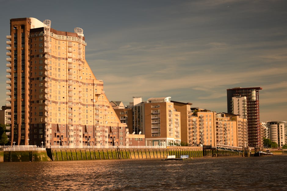 A marina with multiple moored narrowboats and small sailing vessels, anchored along the edge of calm water under a partly cloudy sky. Behind the boats, there are modern residential buildings with balconies and rooftop extensions, alongside taller office or apartment blocks in the distance. The scene depicts the waterfront area in Silvertown, with a clear view of the urban skyline and surrounding structures. The boats include various colors such as green, blue, and red, and are secured with ropes. The water reflects the boats and buildings, creating a mirror-like effect. Nearby, a paved walkway and small dock area are visible, suggesting a location used for boat access and home relocation activities. This setting provides a typical scene for shipping, loading, or preparing for home removals or furniture transport through the area, supported by ongoing moving services like those offered by Man with Van Silvertown.