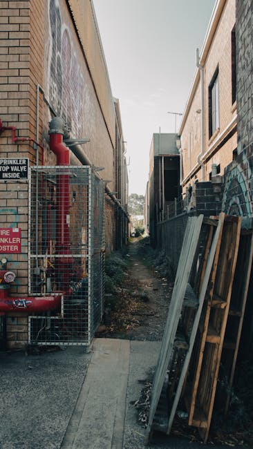 A narrow alleyway between two brick buildings, with construction materials and equipment visible. On the left side, a metal fence encloses fire safety equipment, including red pipes and a fire sprinkler system, with a sign indicating 'Fire Sprinkler' and safety instructions. On the right side of the alley, several wooden pallets and metal panels are leaning against the building, indicating ongoing building work or renovation. The ground is uneven, covered with dirt and debris, and a small concrete path runs through the middle, leading towards a distant view of a partly cloudy sky. This setting reflects a typical preparation or staging area for a home relocation or furniture transport involving outdoor loading and unloading, with the work area being adjacent to the property for a smooth move facilitated by Man with Van Silvertown.