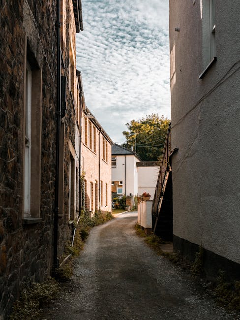 A narrow residential alleyway with a dirt and gravel surface, flanked by older brick and stone houses on the left and a white-painted building on the right. The buildings have small windows, some with shutters, and the structures show signs of age and weathering. In the background, there are trees, a white modern house, and a partly cloudy sky overhead. The scene appears to be in a quiet urban neighbourhood, suitable for home relocation or moving services like those offered by Man with Van Silvertown, which provides removals for tight or narrow street access. The alley is well-lit with natural daylight, emphasizing the textured surfaces of the buildings and pavement, and the area could be used for loading or unloading furniture and packing materials during a move or home packing process.