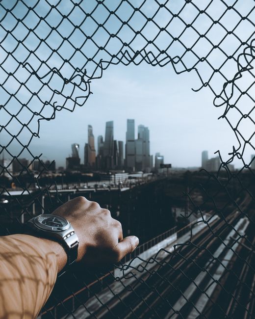 Close-up of a person's hand wearing a silver watch, gripping a rusted metal fence with holes, overlooking a city skyline with tall, modern buildings under a cloudy sky. The fence appears to be part of an outdoor area near a construction site or an industrial zone. The image captures the contrast between the rough texture of the fence and the sleek, reflective surfaces of the skyscrapers in the background. Natural light illuminates the scene, highlighting the details of the hand, watch, and fence, while the distant cityscape provides a blurred yet recognizable urban setting. This image relates to urban relocation or a home move, reflecting moving logistics and the process of transporting belongings through city environments, which aligns with the services offered by Man with Van Silvertown in residential and commercial removals.