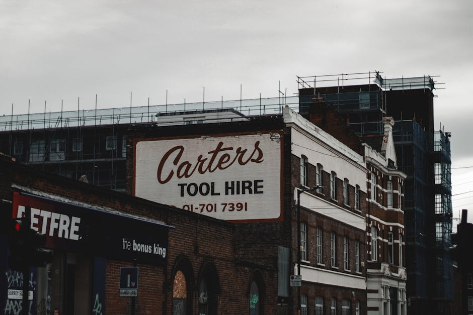 Photograph of a multi-storey building in an urban setting, featuring a large sign for Carters Tool Hire attached to the brick facade. The building is partially covered by scaffolding, indicating ongoing construction or renovation work. In the foreground, a smaller sign for Etfre and The Bonus King is visible, attached to a storefront at street level. The scene is captured during daytime with overcast skies, creating diffused lighting. The architecture includes brickwork and white trim details, typical of building exteriors in city environments. This setting suggests a location where professional house removals or furniture transport may take place, with the infrastructure supporting building upgrades or refurbishments that require careful management of moving and disposal logistics. The image represents a typical urban renewal area where moving services like those offered by Man with Van Silvertown could be involved in home relocation or furniture transport tasks.