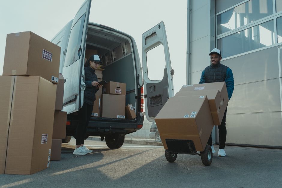 Two movers from Man with Van Silvertown are engaged in a home relocation process outside a modern building, with the loading area on a paved surface. One mover, a woman wearing a black jacket, white s